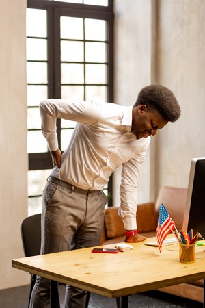 Unpleasant feelings. Unhappy young man standing near the table in the office while suffering from pain in the backの写真素材