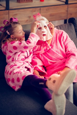 Taking sheet mask. Granny and granddaughter wearing pink bathrobes taking sheet mask while enjoying beauty dayの写真素材