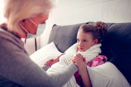 Having flue. Cute dark-eyed girl wearing warm beige scarf while having flue looking at her caring grannyの写真素材