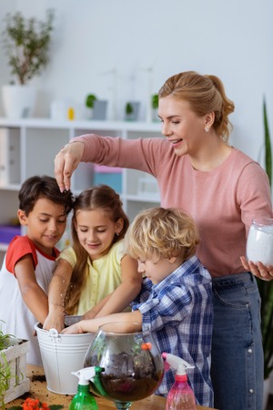Into potting soil. Blonde-haired teacher putting sugar into potting soil near pupils while planting flowers at lessonの写真素材