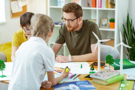 Student asking questions. Concentrated bearded man in clear glasses sitting with his students and drawing posterの写真素材