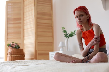 Feeling miserable. Red-haired anorexic woman with tattoos on arms feeling miserable while sitting on...