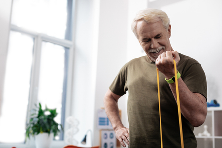 Sports equipment. Joyful strong man stretching his arm with a rubber band while trainingの写真素材
