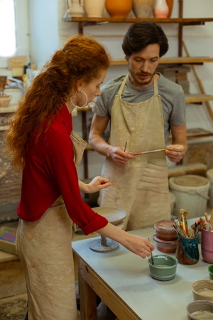 Starting painting process. Smart dark-haired master holding brushes and talking to young student about painting techniquesの写真素材