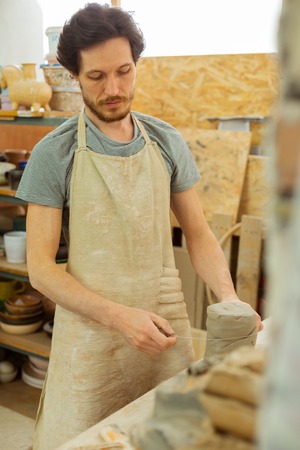 Fresh clay. Focused dark-haired man in special apron measuring needed amount of clay with thread