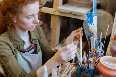 Preparing for creating . Serious curly woman sorting out paintbrushes in big pile while organizing her working processの写真素材