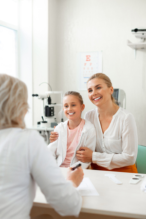 Attentive doctor. Loving mother with wide smile supporting her little girl during medical appointmentの写真素材