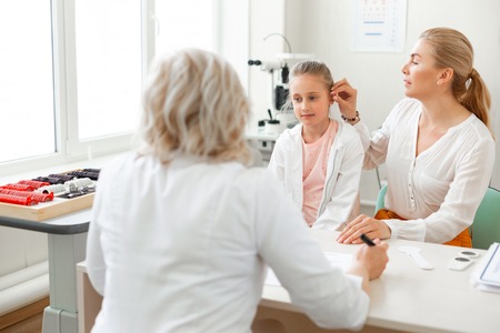 Work with patients. Caring long-haired mother visiting doctor with her daughter and describing their situationの写真素材