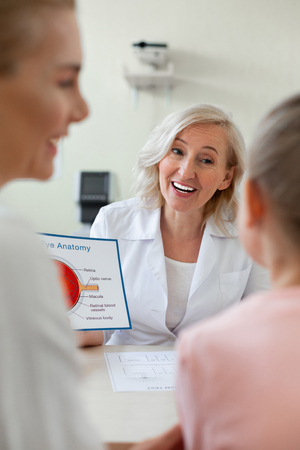 Presenting picture. Cheerful blonde doctor in white robe sitting in front of her patients and talking about eye issuesの写真素材