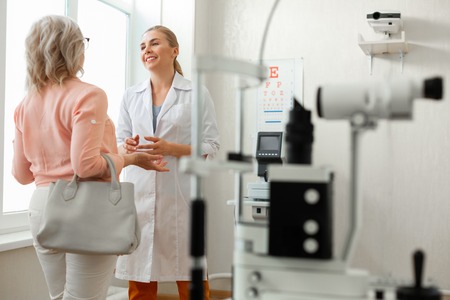 Near bright window. Pleasant kind doctor in long white robe talking to patient while staying in spacious cabinetの写真素材
