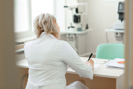 Experienced blonde oculist. Focused short-haired doctor in white robe sitting at the table and writing down recommendationsの写真素材