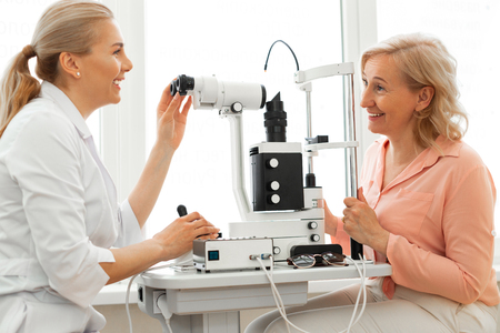 Checking eyesight. Smiling good-looking oculist with tied hair having pleasant appointment with senior patientの写真素材