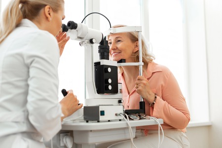 Frame of device. Smiling good-looking senior lady sitting in front of professional oculist with giant device between themの写真素材