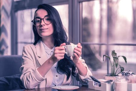 Tasty cappuccino. Delighted nice woman holding a cup of coffee while being in the cafeteriaの写真素材