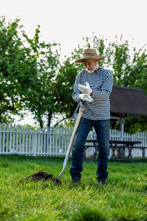 Working with shovel. Grey-haired man wearing gloves working with shovel in garden near cottage house