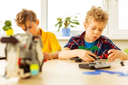 Two classmates. Nice blonde boy sitting with his friend while constructing a toy
