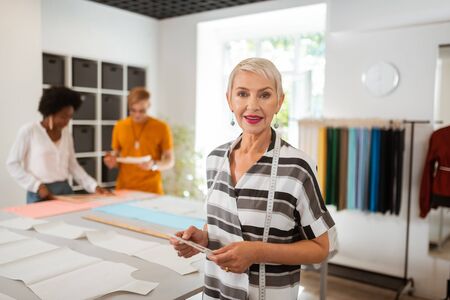 Smiling tailor. Senior female tailor with a measuring tape on the shoulders smiling while standing in front of the cutting tableの写真素材