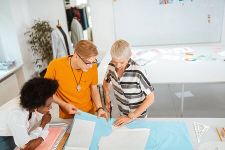 Cutting out patterns. Senior female fashion designer cutting out a pattern while her male colleague...