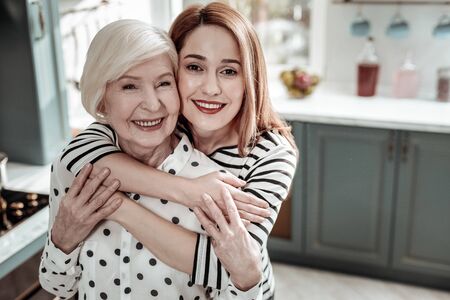 Hugging relatives. Beautiful happy young woman smiling and standing behind her positive senior mother while hugging herの写真素材