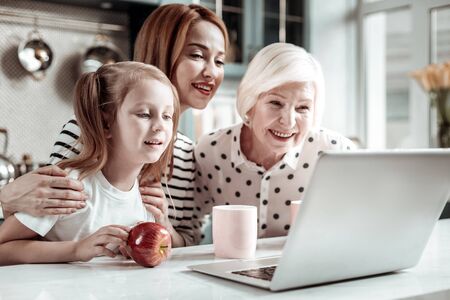 Video call. Progressive women of three generations feeling interested and leaning to the screen of modern laptop while having video callの写真素材