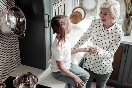 Milk for granddaughter. Cute little girl sitting in the kitchen while her cheerful friendly senior grandmother offering her a glass of milkの写真素材