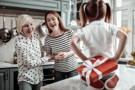 Hidden present. Emotional middle aged woman and her mother looking excited and smiling while cute little girl hiding her beautiful presentの写真素材