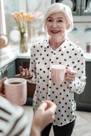 Feeling good. Cheerful beautiful aged woman looking stylish and glad while standing with a cup of tea and having a pleasant conversationの写真素材