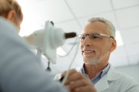 Eye doctor smiling. Bearded grey-haired eye doctor wearing glasses and white uniform smilingの写真素材