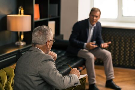 Attentive listener. Back view portrait of a grey-haired psychotherapist sitting in an armchair while making notes and listening to a patientの写真素材