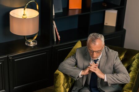 Fingers crossed. Pensive serious grey-haired man sitting with his fingers crossed in an armchair...