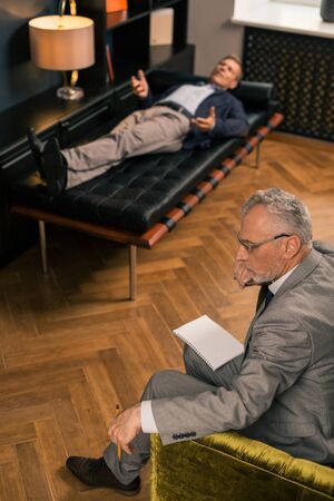Pensive psychotherapist. Side view of a thoughtful serious psychotherapist sitting next to his patient while holding a pencil in his handの写真素材
