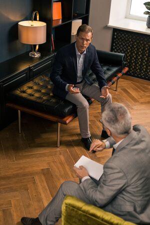 Restless patient. Vertical shot of a restless man sitting on the couch in front of his psychoanalyst while sharing his psychological problemsの写真素材