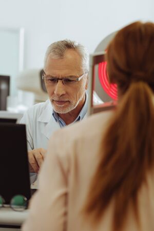 Monitoring eye sight. Grey-haired eye specialist feeling busy while monitoring eye sight of woman