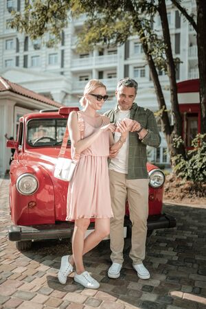 Scrolling together. Cheerful married couple standing outside near their car and using a smartphone...
