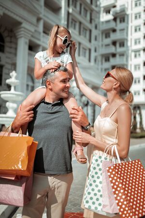 Taking care. Smiling beautiful woman rearranging sunglasses of her daughter sitting on the shoulders of her father after shopping.の写真素材