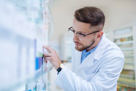 It is here. Handsome brunette chemist standing near shelves and taking package with pillsの写真素材