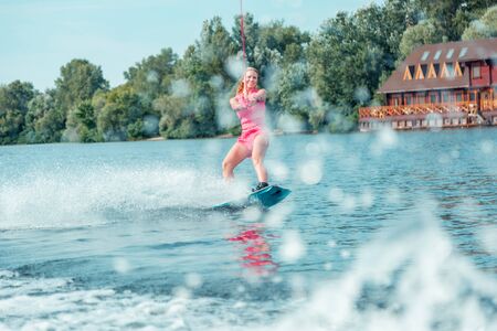 Extreme sport. Smiling blonde long-haired female wakeboarder sliding over the water on a wakeboard in summerの写真素材
