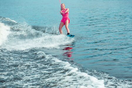 Good mood. Pleased woman wakeboarder in a pink life jacket and shorts smiling while riding a wakeboardの写真素材