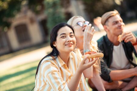 Among your friends. Beautiful smiling girl sitting on the grass and eating pizza spending evening...