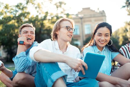 Resting with friends. Three smiling groupmates sitting together with other students on the grass lawn relaxing after classes outside the university.の写真素材
