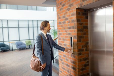 Brown leather bag. Grey-haired mature businessman with brown leather bag calling for elevatorの写真素材
