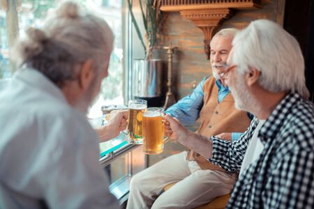 Glasses with beer. Bearded grey-haired men smiling while clanging glasses with beerの写真素材