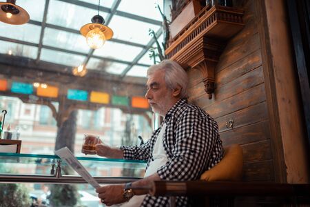 Man reading news. Grey-haired man wearing checked shirt reading news and drinking alcoholの写真素材