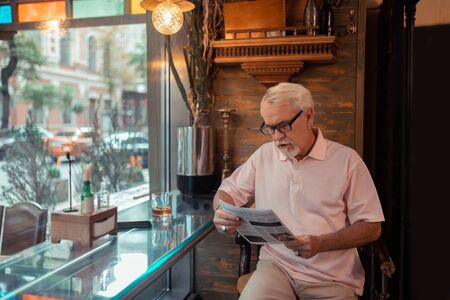 Reading news. Aged grey-haired man wearing pink t-shirt reading news in the pub