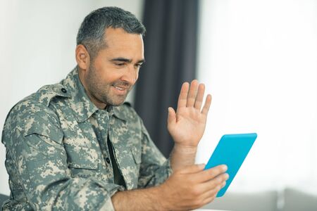 Military man waving. Dark-haired military man waving while having video chat with family on tabletの写真素材