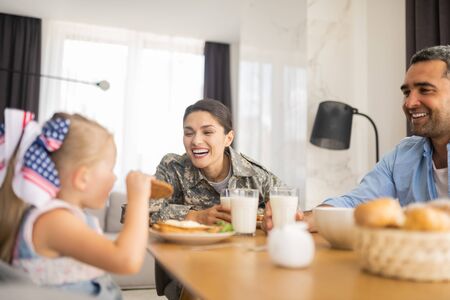 Daughter eating cookie. Appealing happy military woman laughing while looking at her daughter eating cookieの写真素材