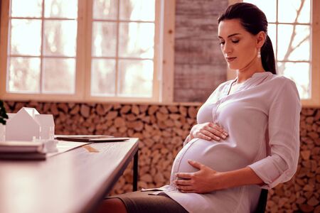 Feel the moment. Charming future mommy sitting at her workplace while massaging her abdomen