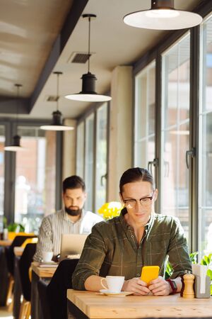 Modern igen. Pleasant young man sitting with his smartphone while using it for online communicationの写真素材