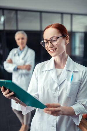 Happy doctor. Smiling good-looking young female doctor looking at a clipboard while standing in the corridor next to her senior colleagueの写真素材