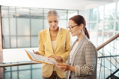 Financial documents. Side view of a young red-haired smiling female entrepreneur looking at documents in the hands of her senior colleagueの写真素材